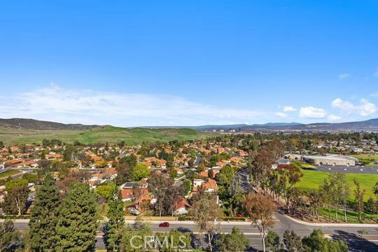 Aerial view of a residential area with houses and trees, set against a background of hills and open sky.