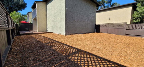 Side view of a two-story house with a wooden fence and mulch-covered yard.
