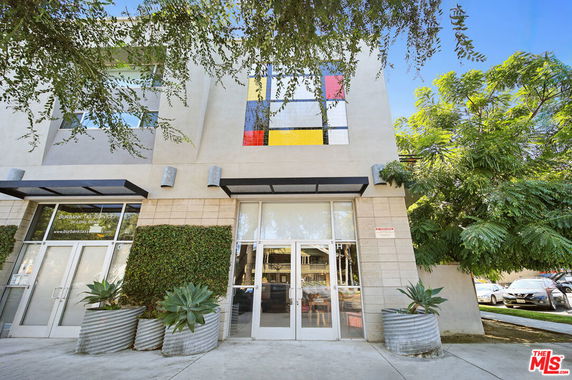 Front view of a modern building with large glass doors and windows, adorned with a colorful geometric design and surrounded by trees and potted plants.