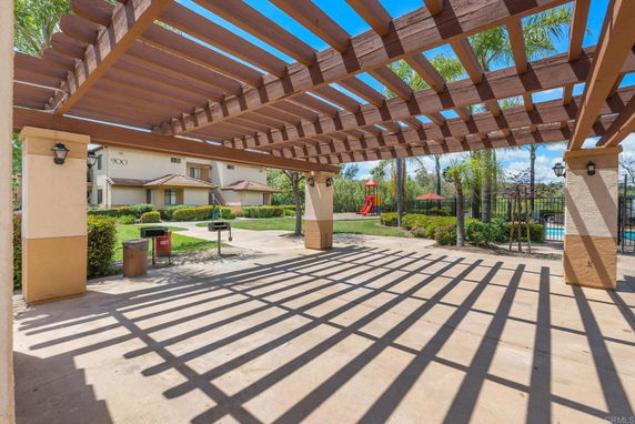 Outdoor area with a pergola structure, adjacent to a playground with slides and green landscaping.
