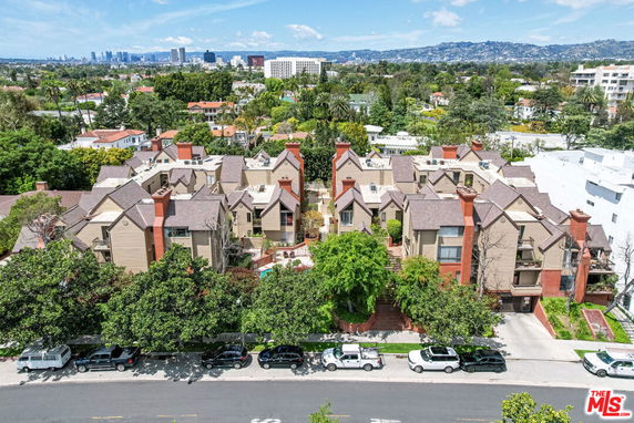 Aerial view of a residential complex with multiple buildings and chimneys, surrounded by a green landscape and urban skyline in the background.