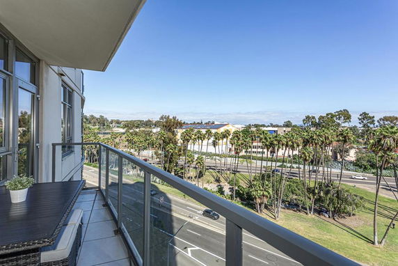 View from a balcony overlooking a street lined with tall palm trees and a distant building under a clear blue sky.