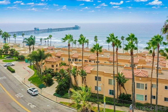 A coastal residential building near the beach with a visible pier and palm trees.