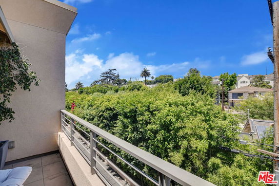View from a balcony showing lush green trees and distant houses under a clear blue sky.