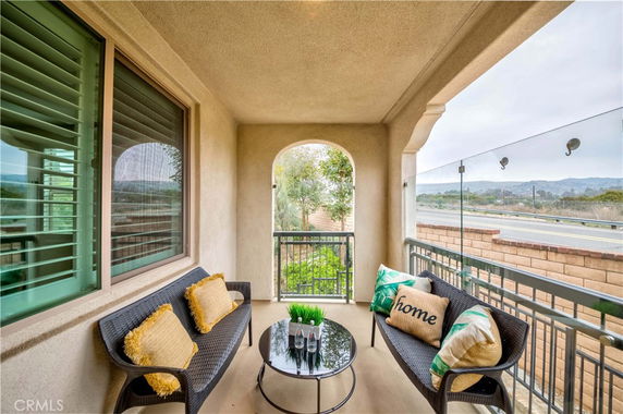 Balcony view from a house featuring glass railing, outdoor seating, and overlooking a scenic road and hills.