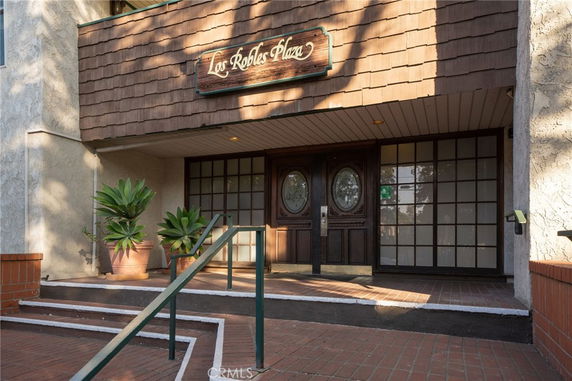 Front view of a building entrance with a wooden sign reading 'Los Robles Plaza', featuring double doors and potted plants.