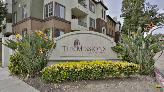 Front view of an apartment building with a visible entrance sign and surrounding greenery.