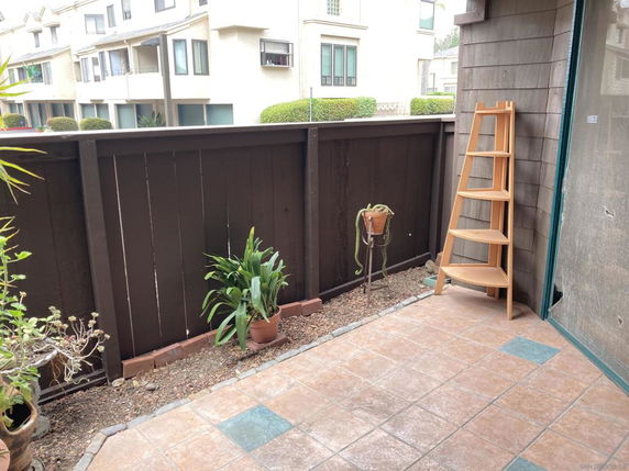 View from a small tiled patio with potted plants and a shelf, overlooking neighboring buildings.