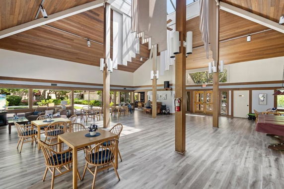 Spacious indoor clubhouse with wooden ceilings and large windows, featuring tables and chairs for recreational activities.