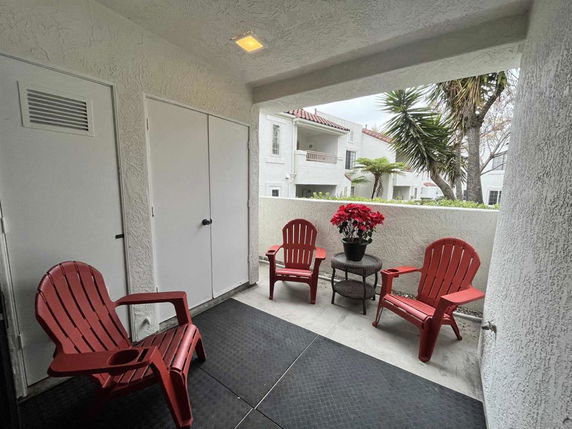 View from a covered patio area with red chairs and potted plant overlooking adjacent buildings and trees.