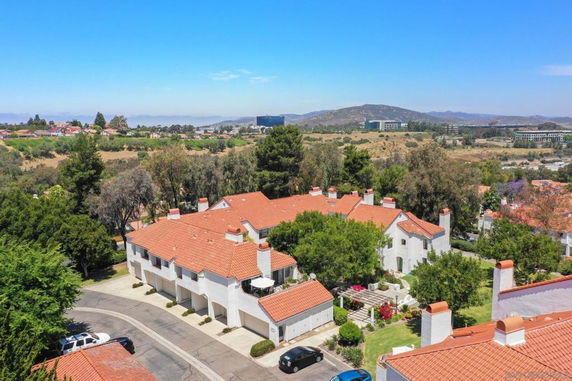 Aerial view of a residential area with white buildings and red tile roofs, surrounded by trees and open landscape.
