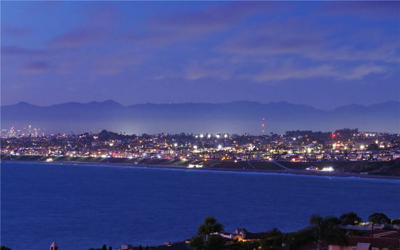Panoramic view of a coastal city at night with lights and mountains in the background.