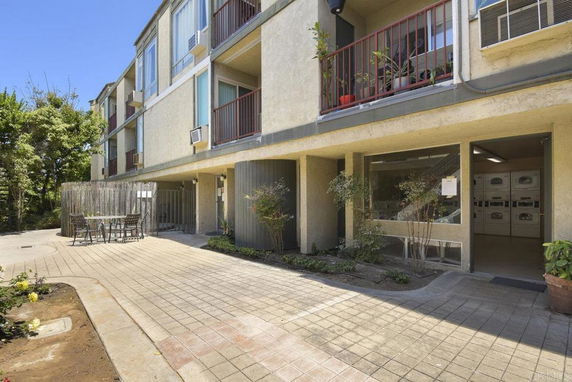 Front view of a multi-story apartment building with balconies and a small outdoor seating area.