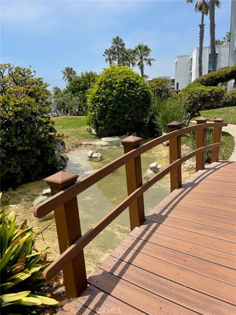 View of a wooden bridge over a small stream with surrounding greenery and a building in the background.