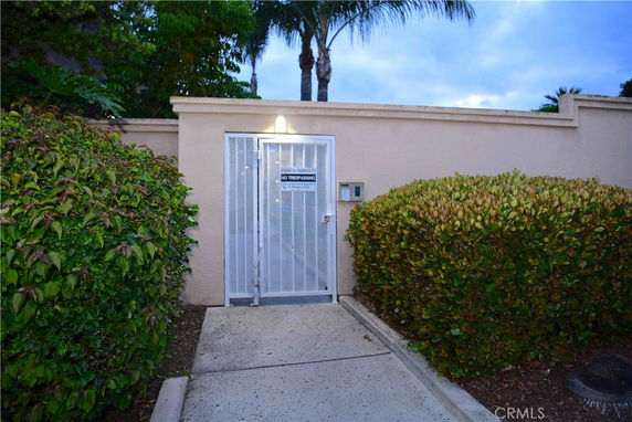 Front view of an entrance gate with metal bars and a no trespassing sign, surrounded by a low wall and bushes.