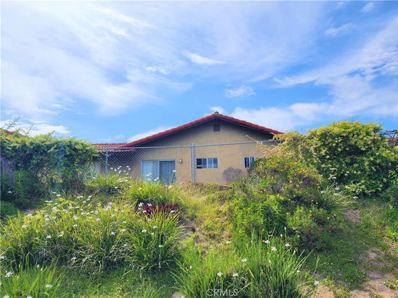 Rear view of a single-story house with a red-tiled roof and beige exterior walls, surrounded by a chain-link fence and overgrown vegetation.