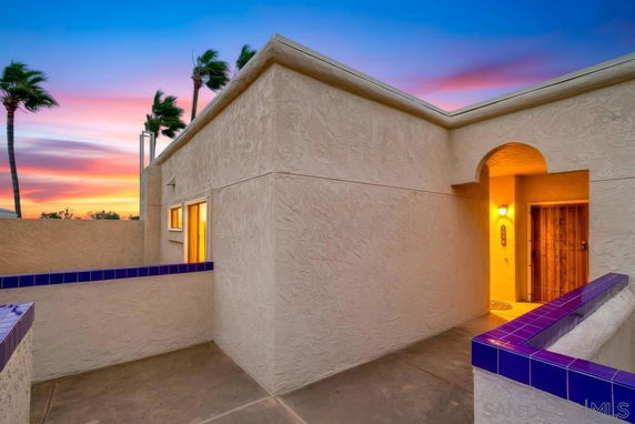 Exterior view of a building with a textured wall and a lit entrance, featuring blue-tiled accents against a sunset sky.