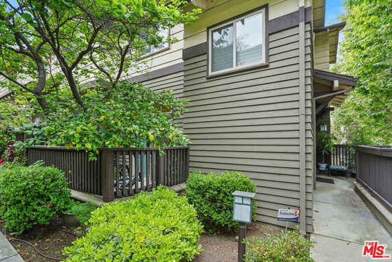 Corner view of a building with wooden siding and a small fenced patio surrounded by greenery.