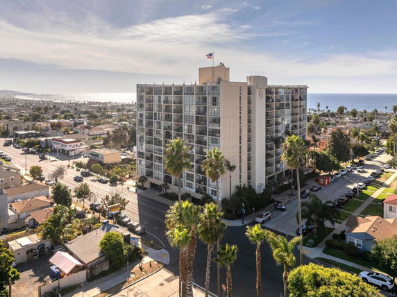 Front view of a tall residential building with balconies and a flag on top, situated in a suburban area near the coastline.