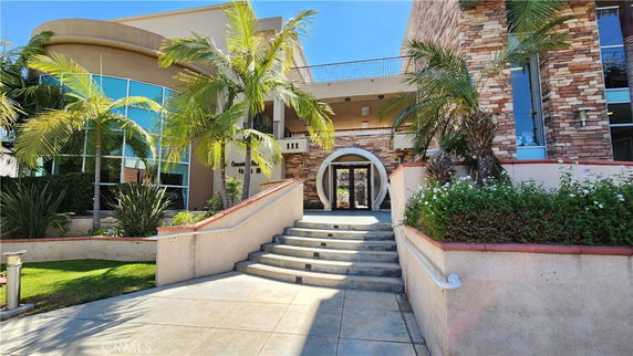 Front view of a modern building with stone and glass facade, featuring palm trees and a curved staircase leading to an entrance.