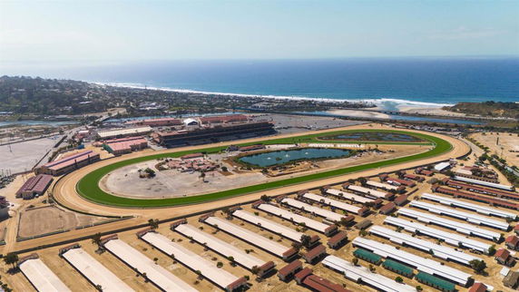 Aerial panoramic view of a large outdoor racetrack and surrounding buildings with the ocean in the background.