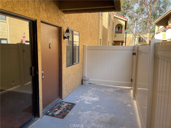 View of the entrance to a unit in a multi-story housing complex with a brown door and white fence.