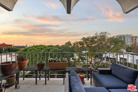 Panoramic view from a balcony with outdoor seating and potted plants overlooking a sunset and surrounding greenery.