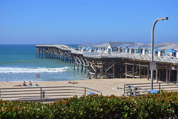 View of beach cottages on a pier extending into the ocean with people relaxing on the beach.