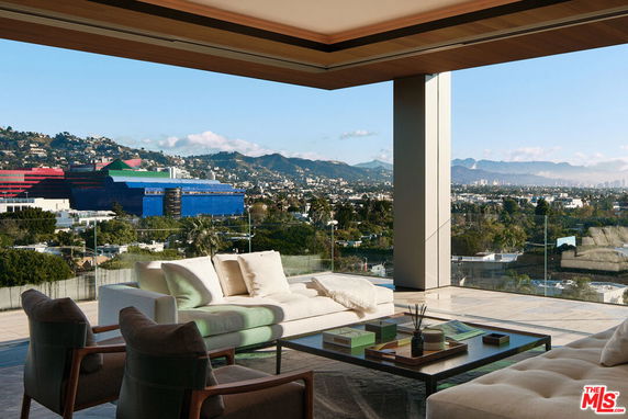 Panoramic view from a modern building showcasing a spacious balcony with luxurious seating and a scenic cityscape with hills in the background.