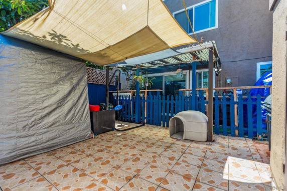 Outdoor area with tiled flooring, covered by a shade, adjacent to a building with a blue picket fence.