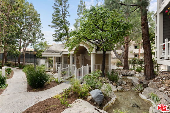 Front view of a small building with a gabled roof and railing, surrounded by trees and landscaped paths.