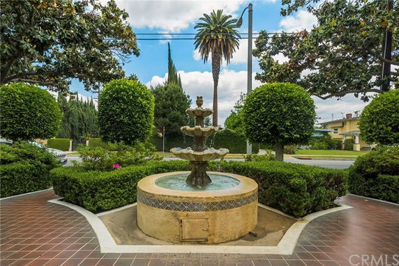 A decorative fountain surrounded by greenery on a tiled patio with a road and palm trees in the background.