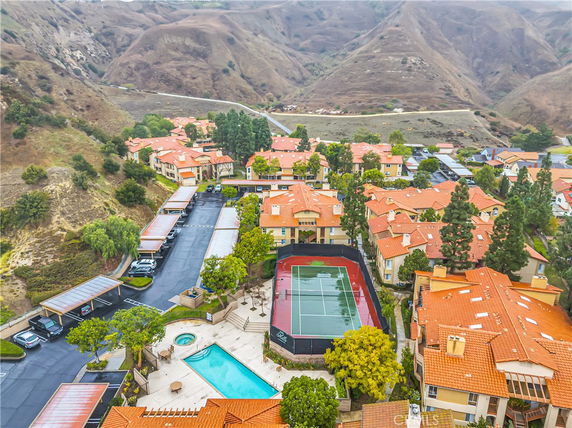 Wide-angle view of a residential area with orange-roofed buildings, a swimming pool, and a tennis court surrounded by hilly landscape.