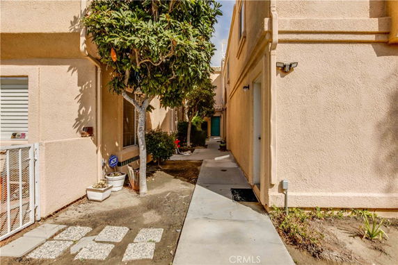 Side view of a beige stucco building with a pathway lined by plants and a small tree.