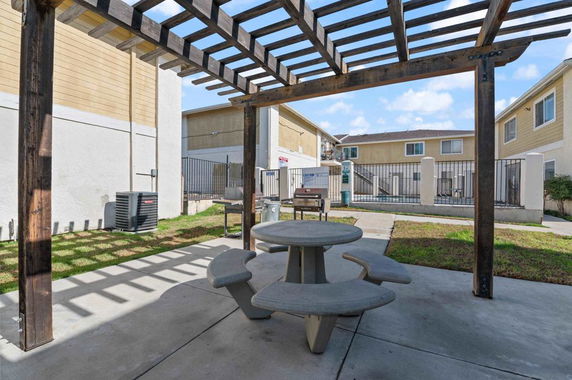 Outdoor area with a round picnic table under a wooden pergola near residential buildings.