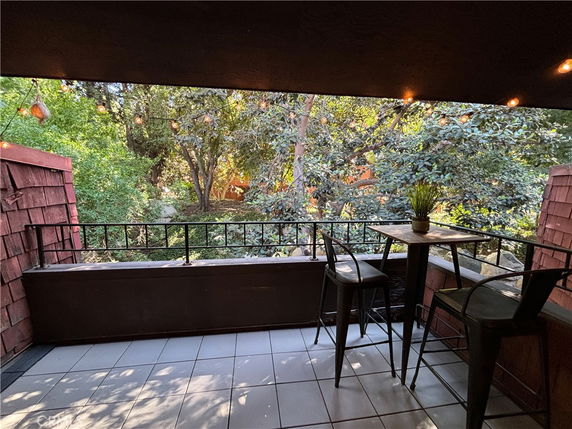 Covered balcony with tiled floor and outdoor furniture, overlooking trees.