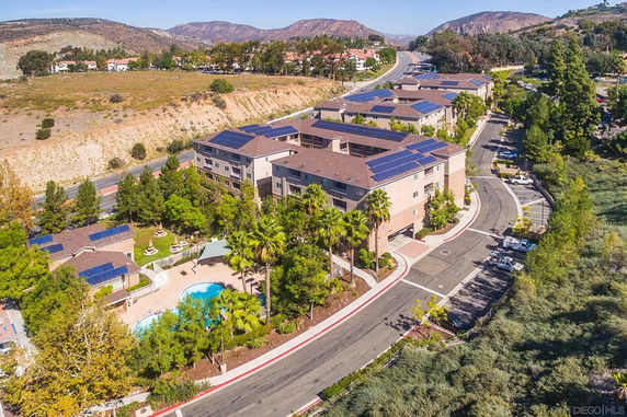 Aerial view of an apartment complex with solar panels on the roofs and a swimming pool, surrounded by trees and hills.