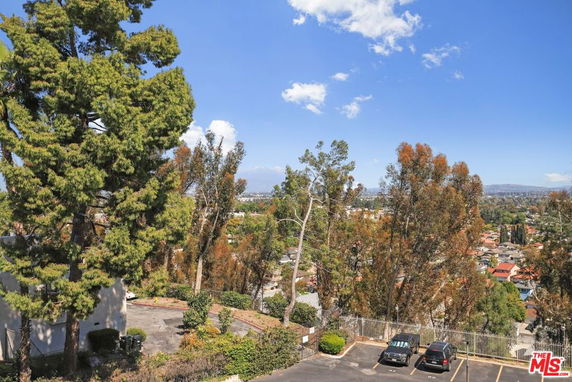 Panoramic view showing trees and a parking area with cars, overlooking a distant cityscape under a clear blue sky.
