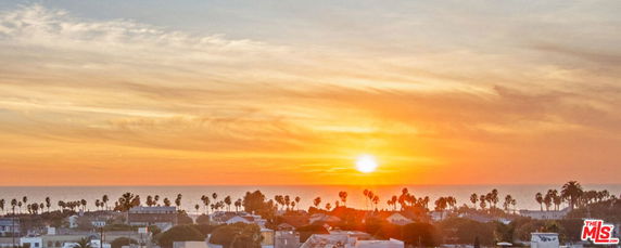 Panoramic view of a sunset over the ocean with silhouetted palm trees and rooftops.