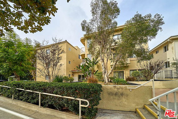 Front view of a multi-story apartment building with beige exterior and surrounding greenery.
