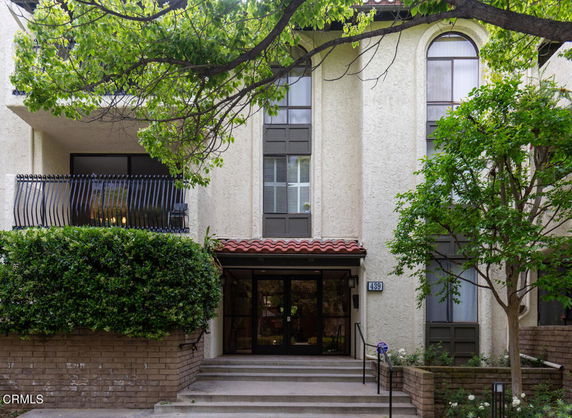 Front view of a building with large windows and a balcony, surrounded by trees and greenery.