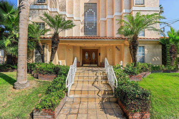 Front view of a multi-story building with an entryway and stairs leading up to the door, surrounded by plants.
