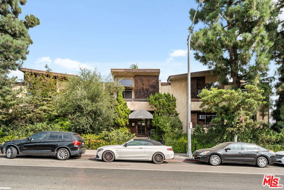 Front view of a two-story residential building with a flat roof and balconies, surrounded by trees.