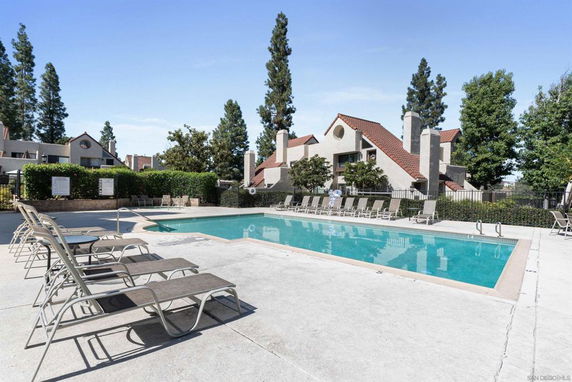 Outdoor swimming pool area with lounge chairs in front of residential buildings with red tiled roofs.