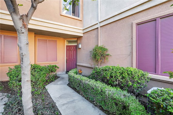 Front view of a house with a red door and pink window shutters, surrounded by small bushes and a tree.