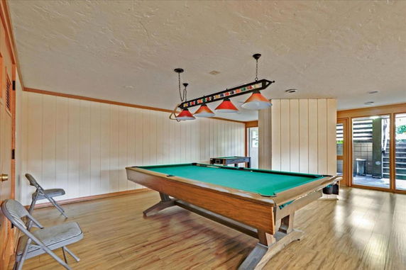 Room with a billiards table and chairs, featuring wooden flooring and paneling, with glass doors leading outside.