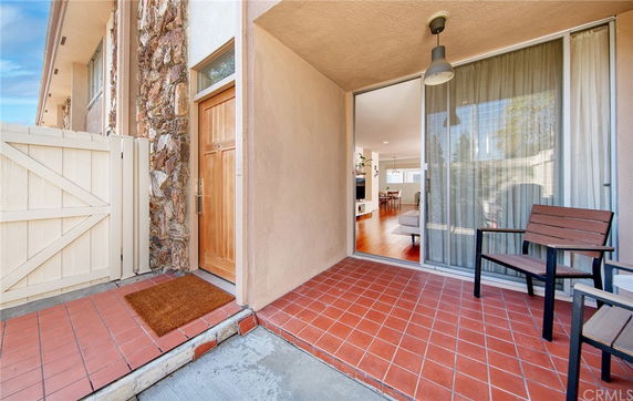 Front view of a house entrance with a wooden door and sliding glass window, featuring a tiled patio area and a wooden chair.
