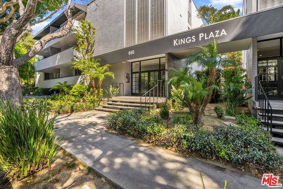Front view of a modern multi-story building labeled 'Kings Plaza' with glass doors and surrounding plants.