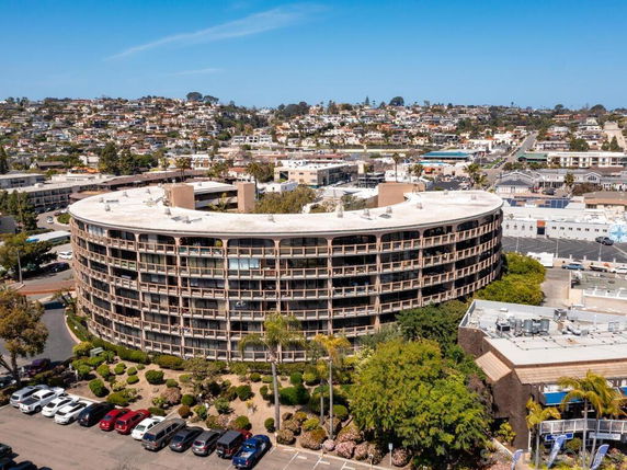 Curved multi-story building with numerous balconies and a parking lot in front.