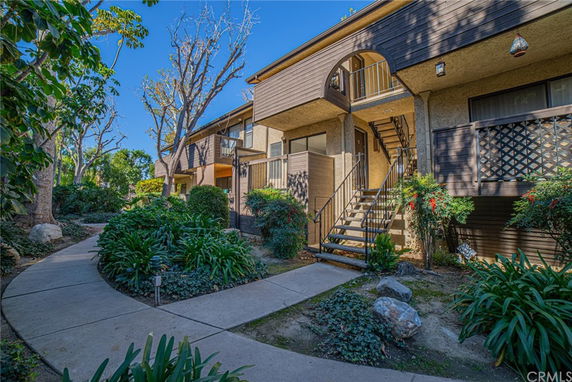 Front view of a two-story building with external staircase and surrounded by leafy green plants.